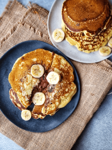 Close-up of a stack of pancakes on a blue plate, oozing with Nutella filling and garnished with fresh banana slices. One pancake has a bite taken out, revealing the chocolatey center.