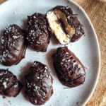Overhead shot of five dark chocolate-covered coconut bars, sprinkled with coconut flakes, on a white speckled plate with a burlap background; one bar is halved, revealing a light-colored coconut filling.