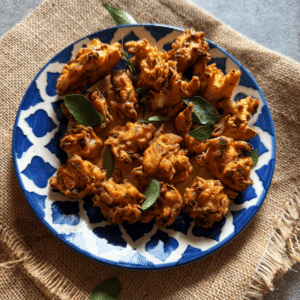 Crispy cabbage pakoras arranged on a blue and white patterned plate, garnished with fresh curry leaves, placed on a textured brown cloth.