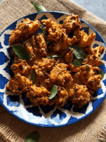 Crispy cabbage pakoras arranged on a blue and white patterned plate, garnished with fresh curry leaves, placed on a textured brown cloth.