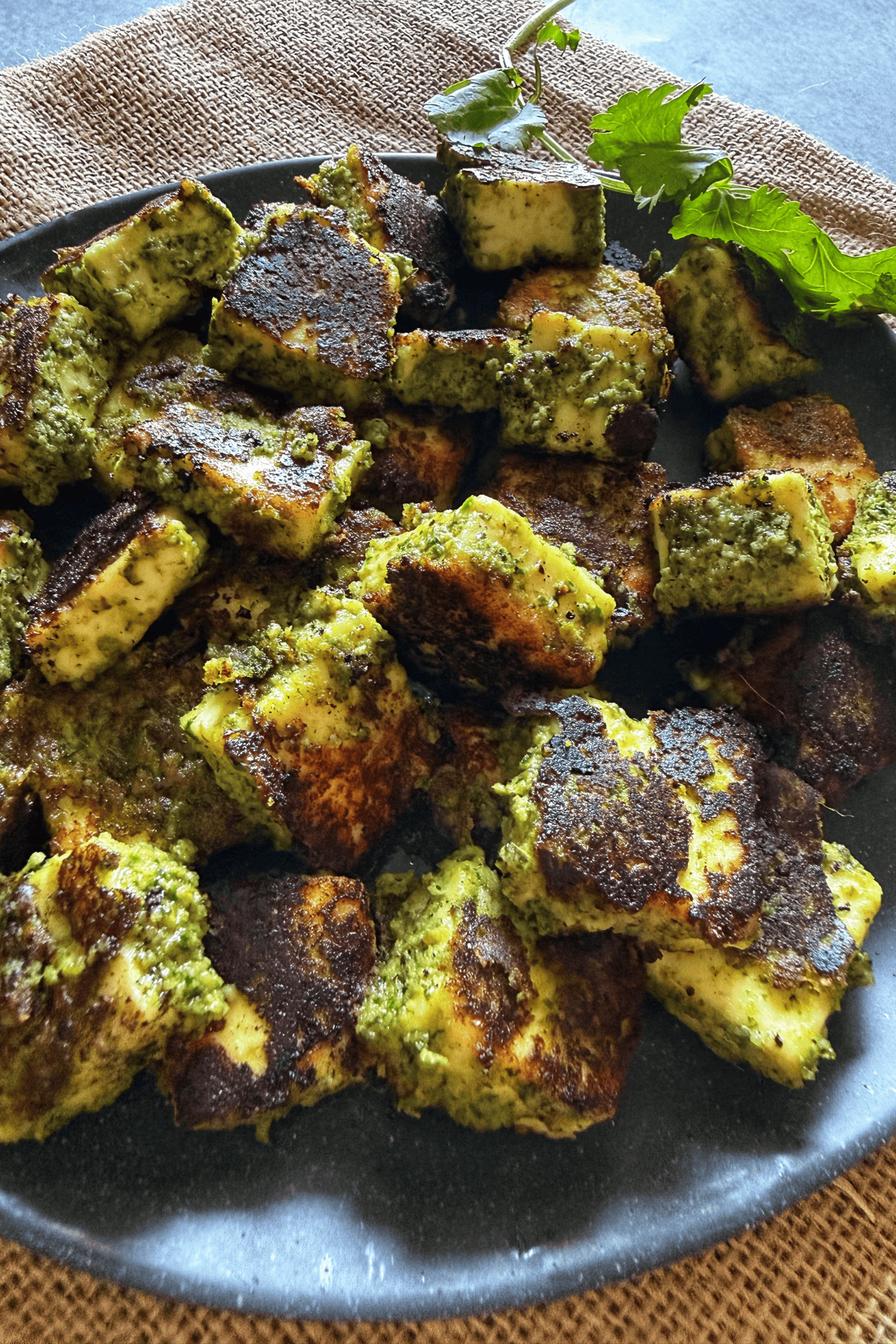 Grilled paneer cubes coated in a vibrant green chutney marinade, served on a black plate and garnished with fresh coriander leaves.