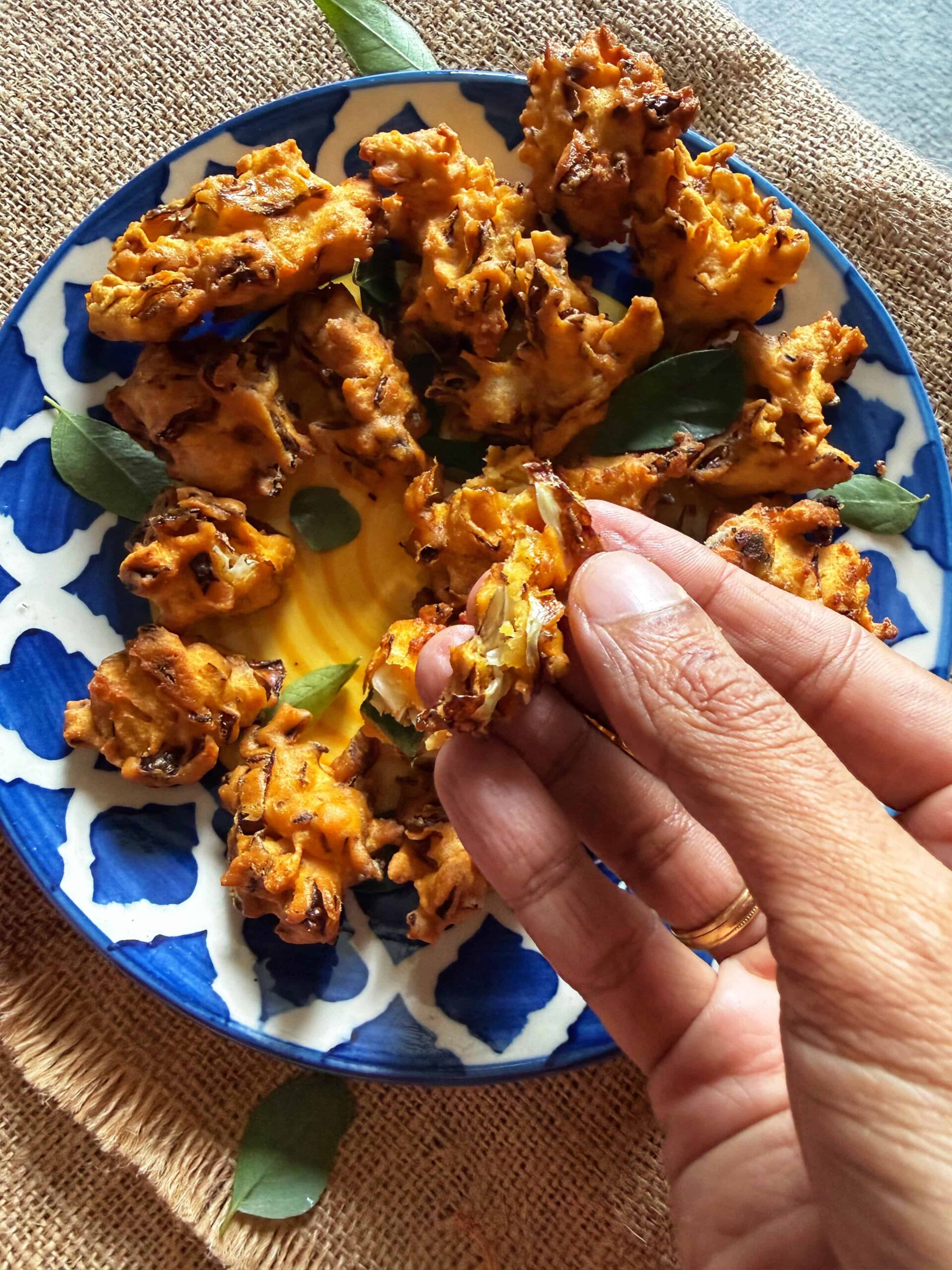 A close-up of a hand holding a piece of crispy, golden-brown cabbage pakora, with several more pakoras served on a blue and white patterned plate garnished with curry leaves, resting on a burlap mat.