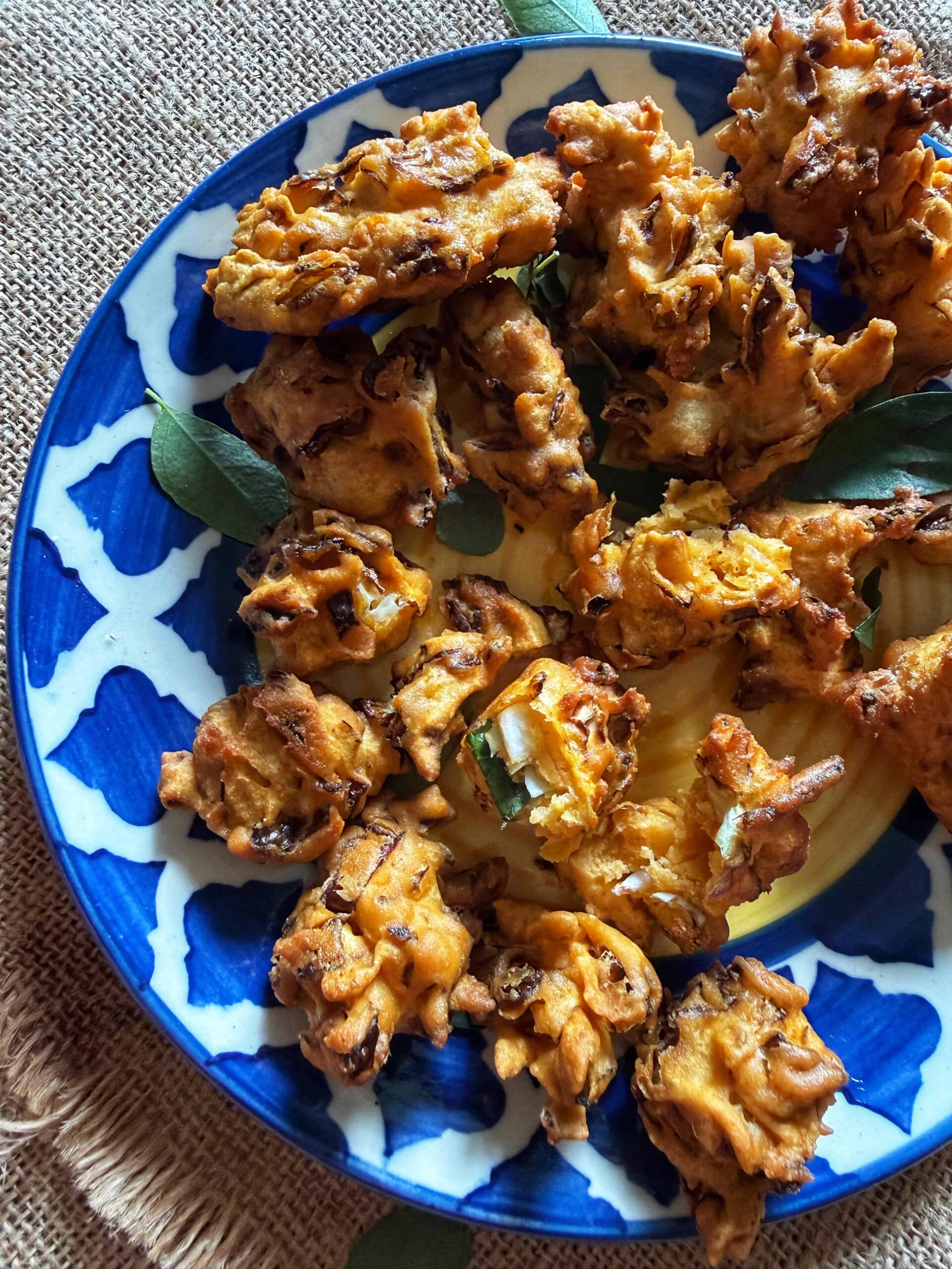 Close-up of crispy cabbage pakoras on a blue and white patterned plate, garnished with curry leaves and placed on a textured burlap cloth.