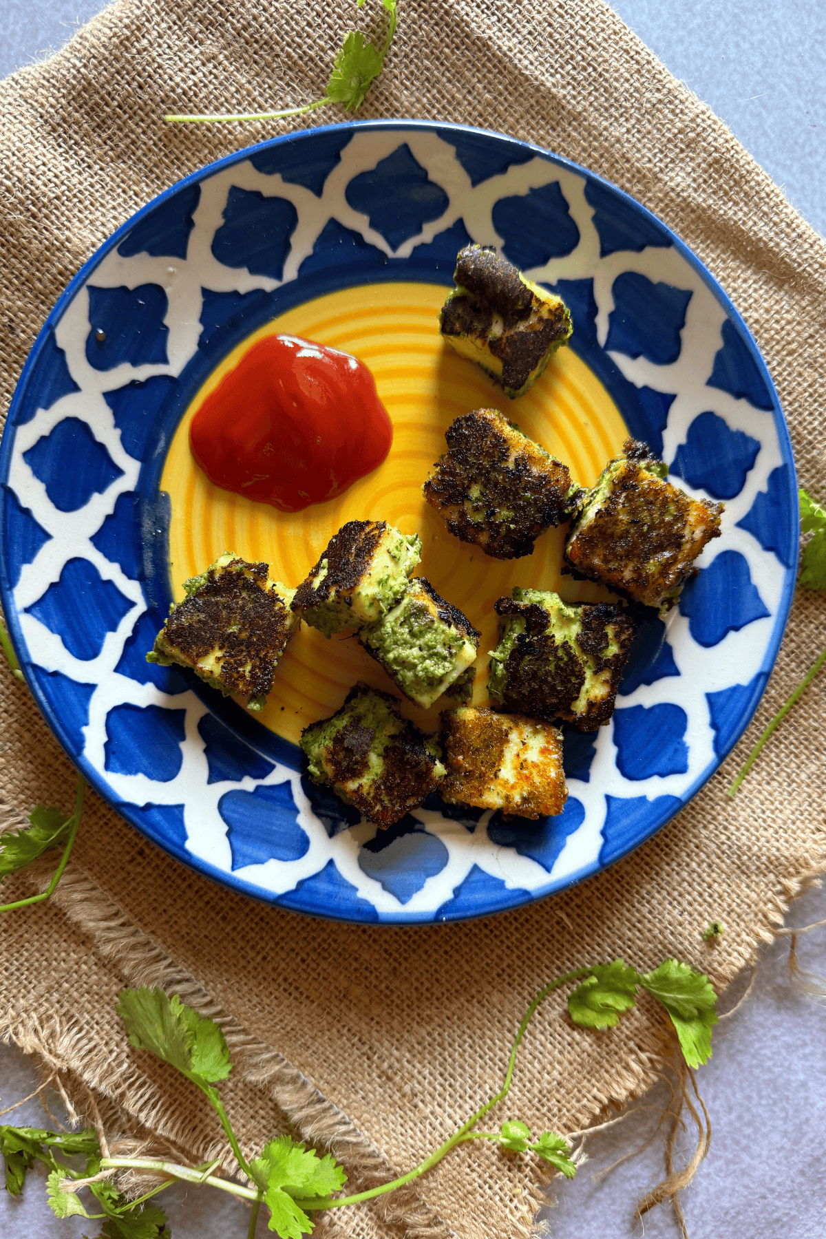 Pan fried paneer cubes coated with coriander peanut paste served on a decorative blue and yellow plate with a dollop of ketchup, garnished with fresh coriander leaves on a burlap cloth background.