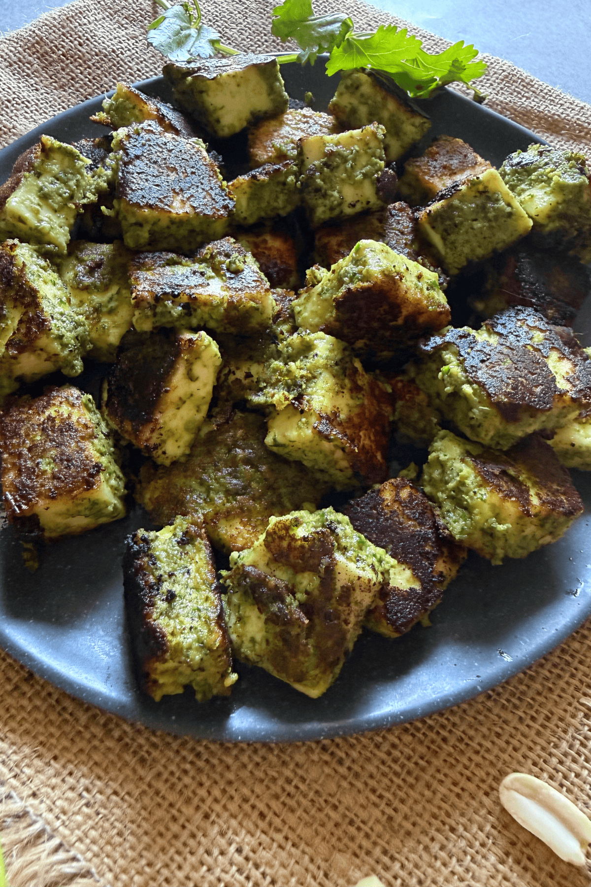 Cubes of pan-fried paneer coated with a vibrant green thecha chutney, served on a dark plate, garnished with fresh coriander on a rustic jute cloth background.
