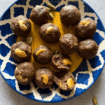 A decorative blue and yellow plate filled with ragi ladoos topped with cashew nuts, surrounded by scattered peanuts and cardamom pods on a light background.