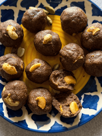 A decorative blue and yellow plate filled with ragi ladoos topped with cashew nuts, surrounded by scattered peanuts and cardamom pods on a light background.