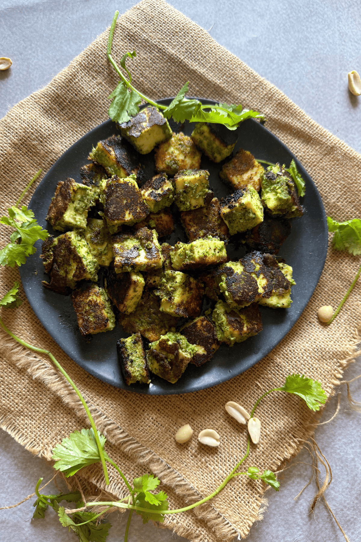 Plated cubes of green-marinated Thecha Paneer, charred and served on a black plate, garnished with fresh coriander leaves and raw peanuts, placed over a rustic burlap mat.