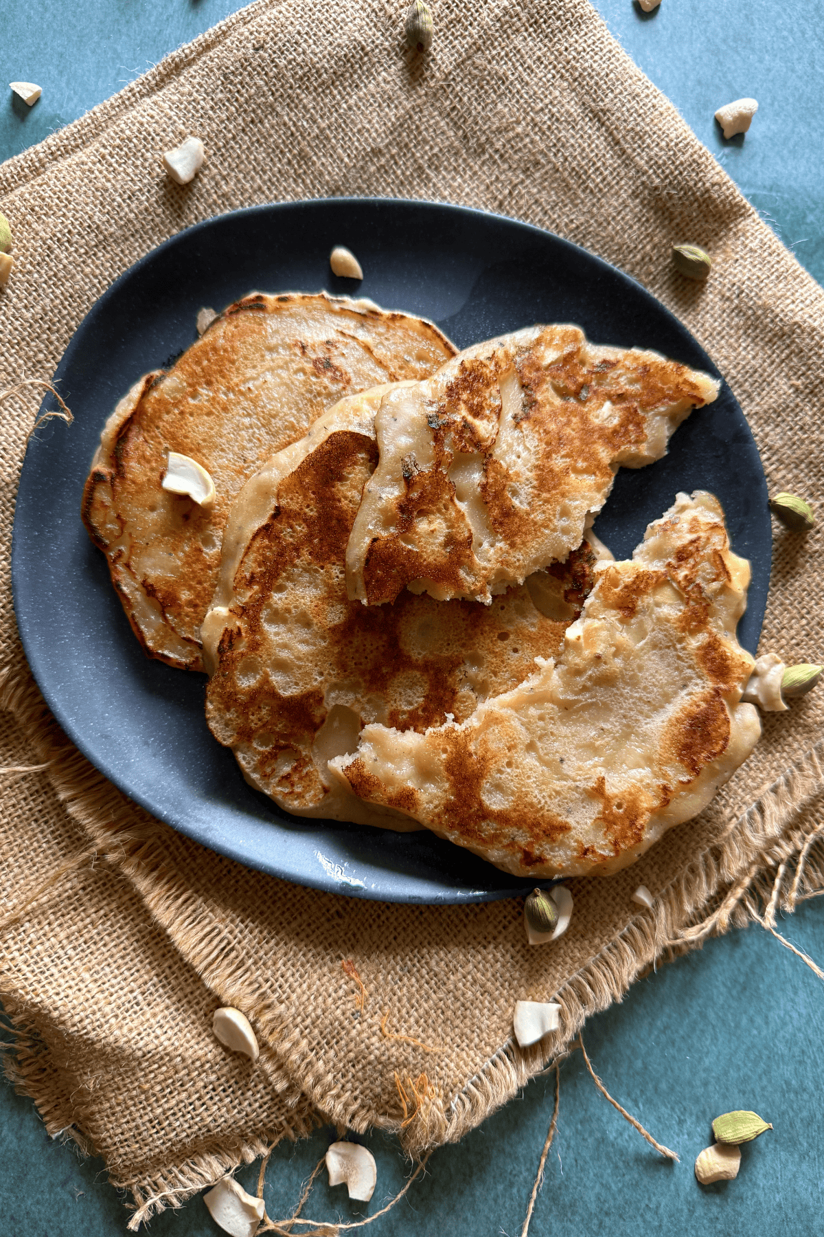 Close-up of three golden-brown banana dosas served on a dark plate, placed over a rustic burlap cloth. The dosas have a slightly crisp, uneven surface and are garnished with scattered cashew pieces and cardamom pods around the plate.