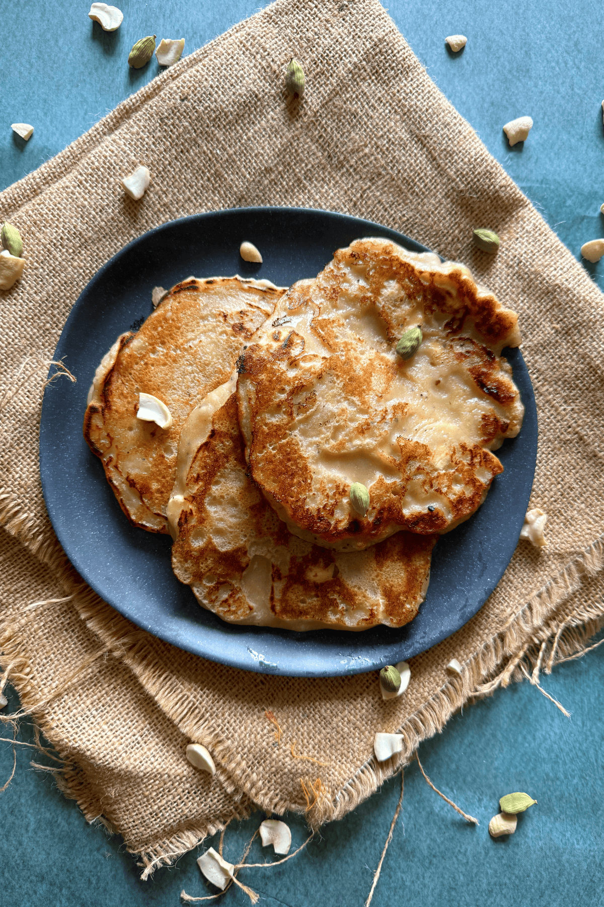 Overhead shot of three golden brown eggless banana dosas on a dark plate, garnished with sliced cashews and cardamom, placed on rustic burlap over a teal background.