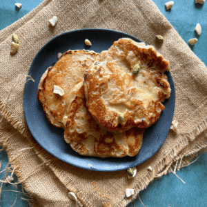 A black plate filled with golden-brown banana dosas, garnished with cashews and cardamom, placed on a rustic burlap cloth with scattered nuts around.