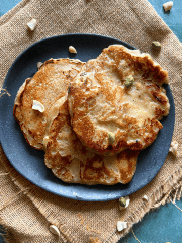 A black plate filled with golden-brown banana dosas, garnished with cashews and cardamom, placed on a rustic burlap cloth with scattered nuts around.