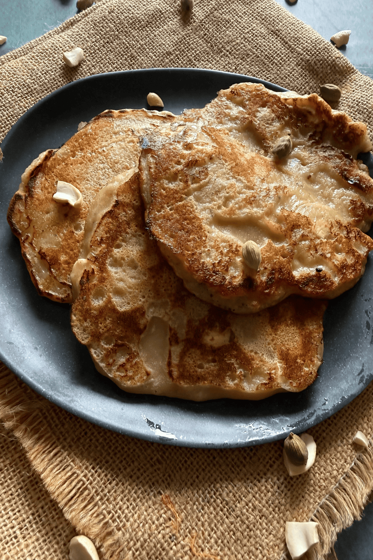 Close-up of three golden-brown banana dosas served on a dark plate, placed over a textured burlap cloth. The dosas have a slightly crispy, uneven surface and are garnished with scattered cashew pieces and cardamom pods.