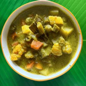A bowl of South Indian Nilgiri vegetable kurma, featuring colorful pieces of carrot, potato, cauliflower, beans, and peas, all cooked in a rich green coconut-coriander masala gravy, served on a green banana leaf background.