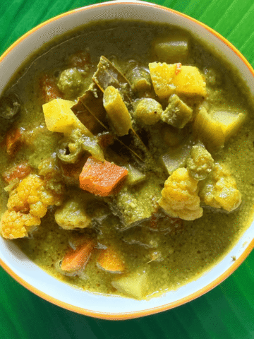 A bowl of South Indian Nilgiri vegetable kurma, featuring colorful pieces of carrot, potato, cauliflower, beans, and peas, all cooked in a rich green coconut-coriander masala gravy, served on a green banana leaf background.