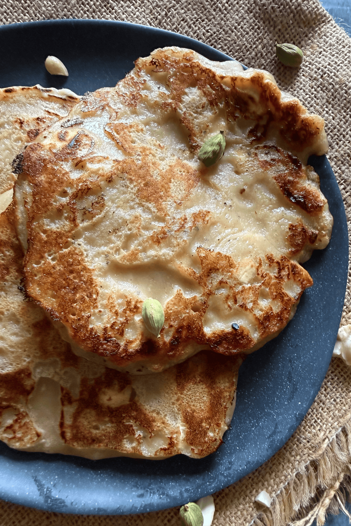 Close-up of golden brown banana dosa topped with whole green cardamom pods on a dark plate over a rustic burlap background.