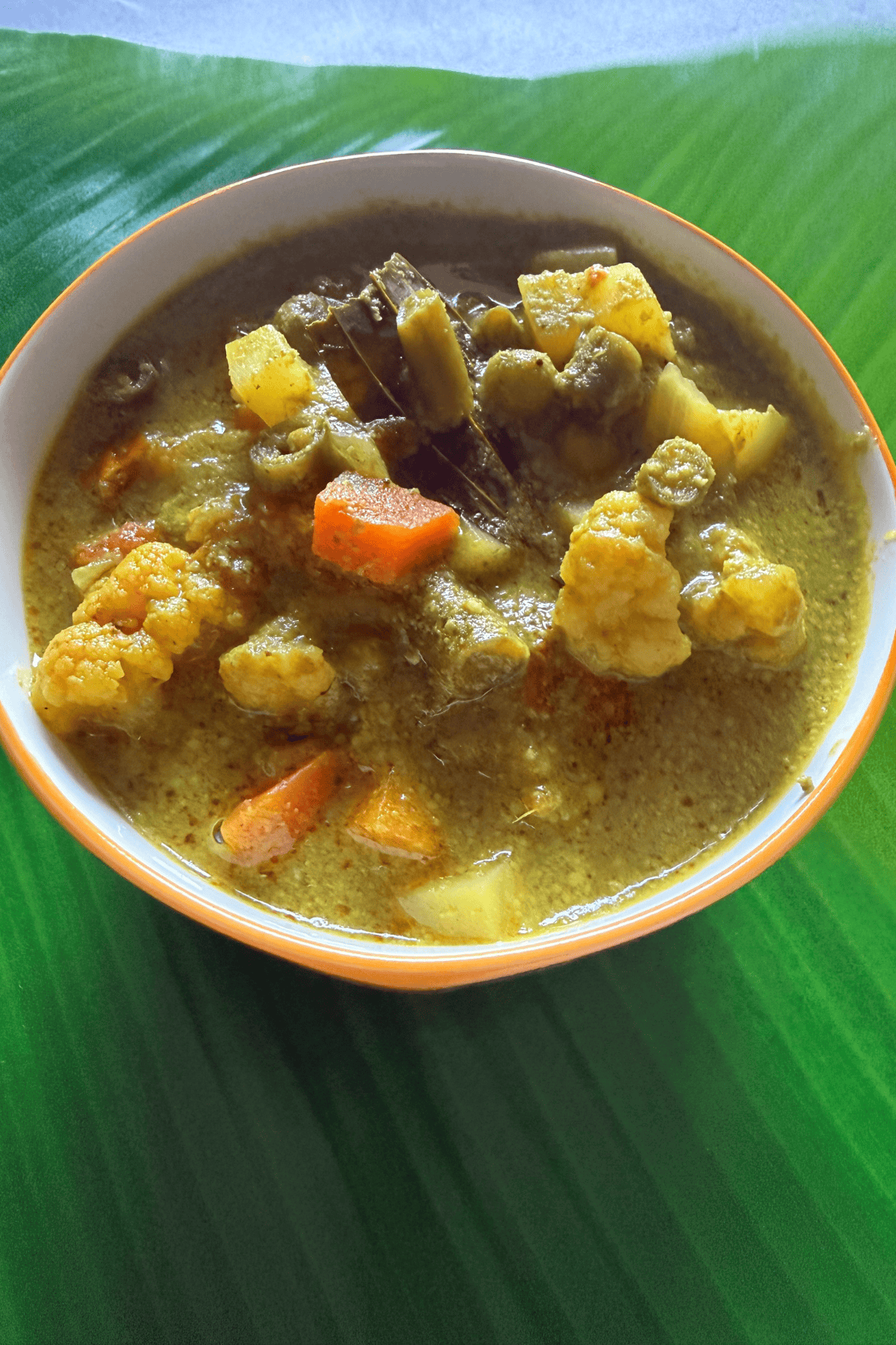 A bowl of South Indian green vegetable kurma featuring carrots, cauliflower, potatoes, and beans in a spiced coconut and herb gravy, served on a banana leaf background.