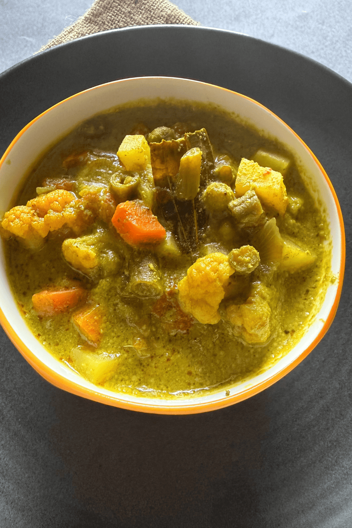 A bowl of South Indian-style green vegetable kurma, featuring mixed vegetables like carrots, cauliflower, beans, and potatoes in a creamy green masala gravy, served in a white bowl with an orange rim on a dark plate.