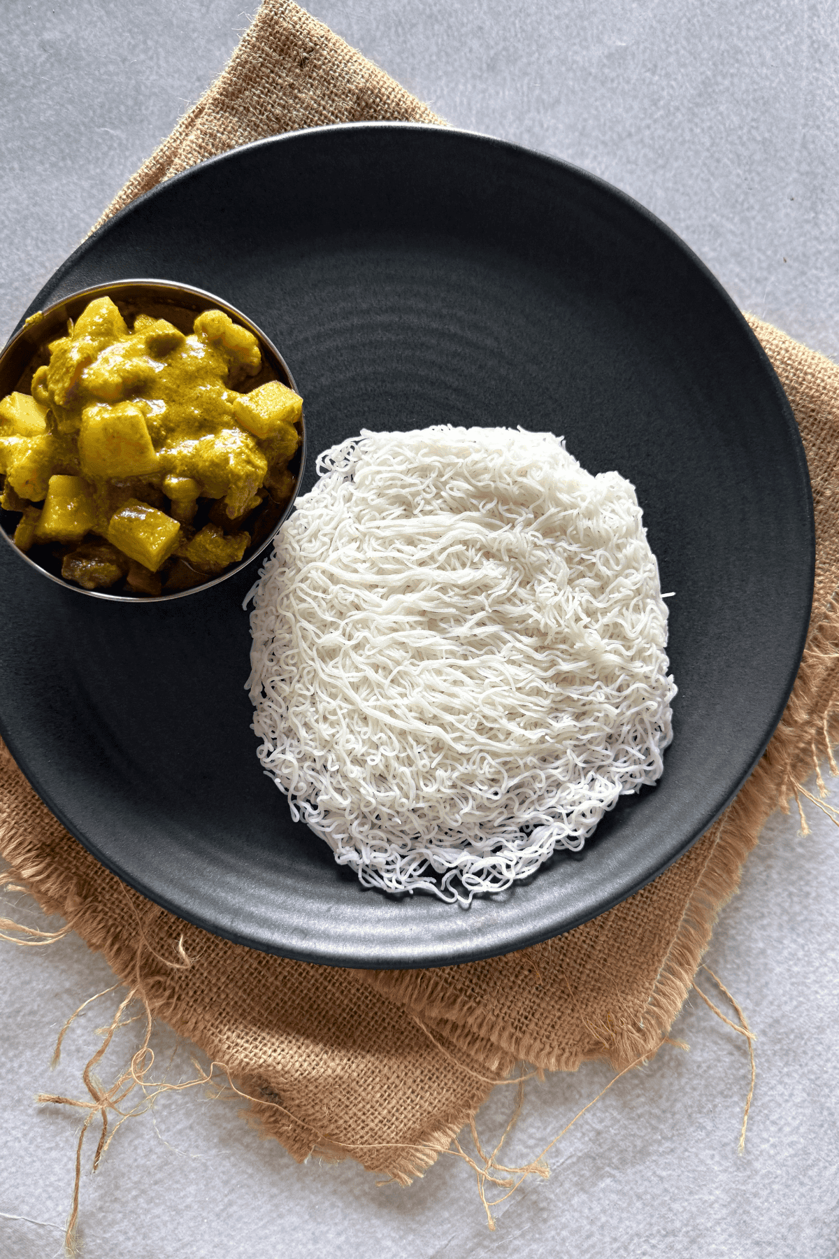 A black plate contains a serving of soft, white idiyappam (string hoppers) with a bowl of green vegetable kurma placed beside it, set on a rustic brown burlap cloth over a light background.