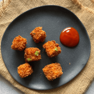 Crispy paneer popcorn arranged on a black plate with a side of tomato ketchup, lightly garnished and set on a rustic burlap cloth.
