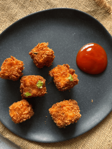 Crispy paneer popcorn arranged on a black plate with a side of tomato ketchup, lightly garnished and set on a rustic burlap cloth.