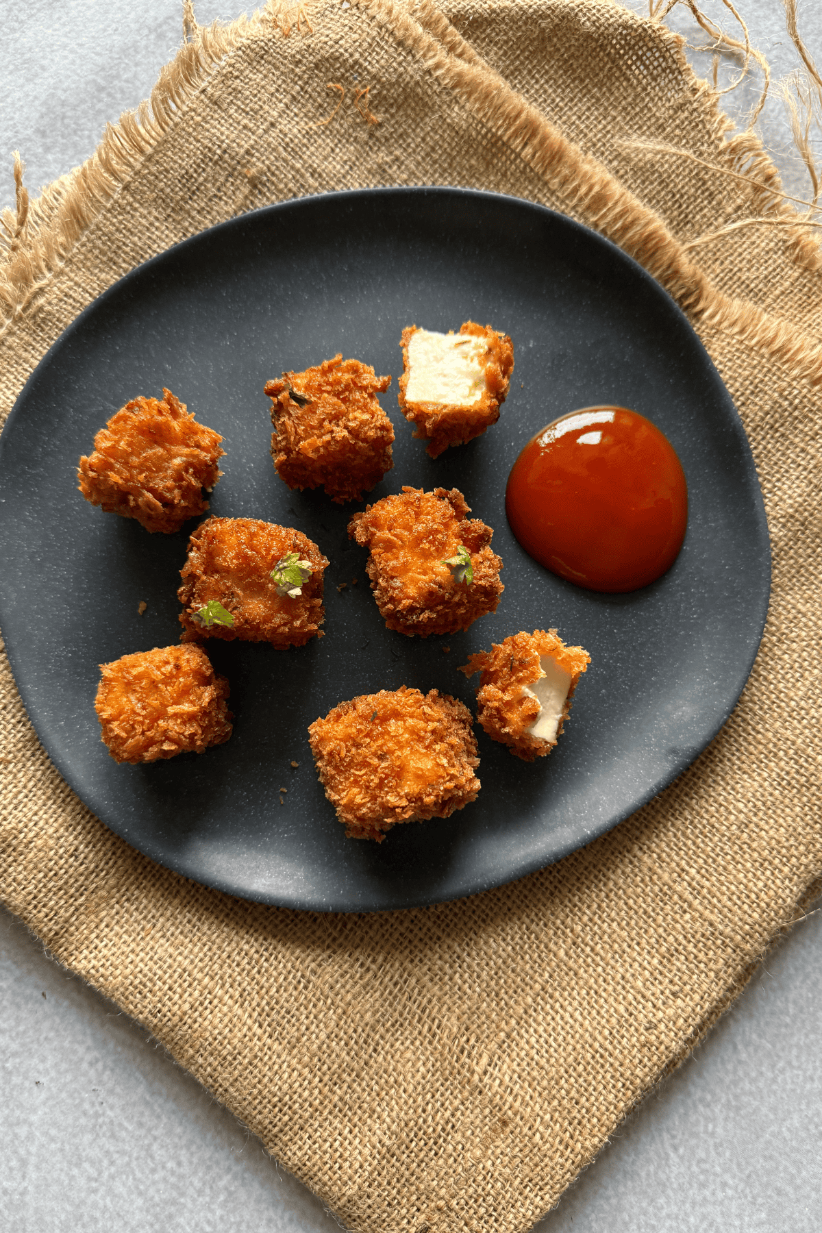 Golden, crispy paneer popcorn served on a black plate with tomato ketchup, one piece broken open to show the soft paneer inside, set on a rustic burlap cloth.