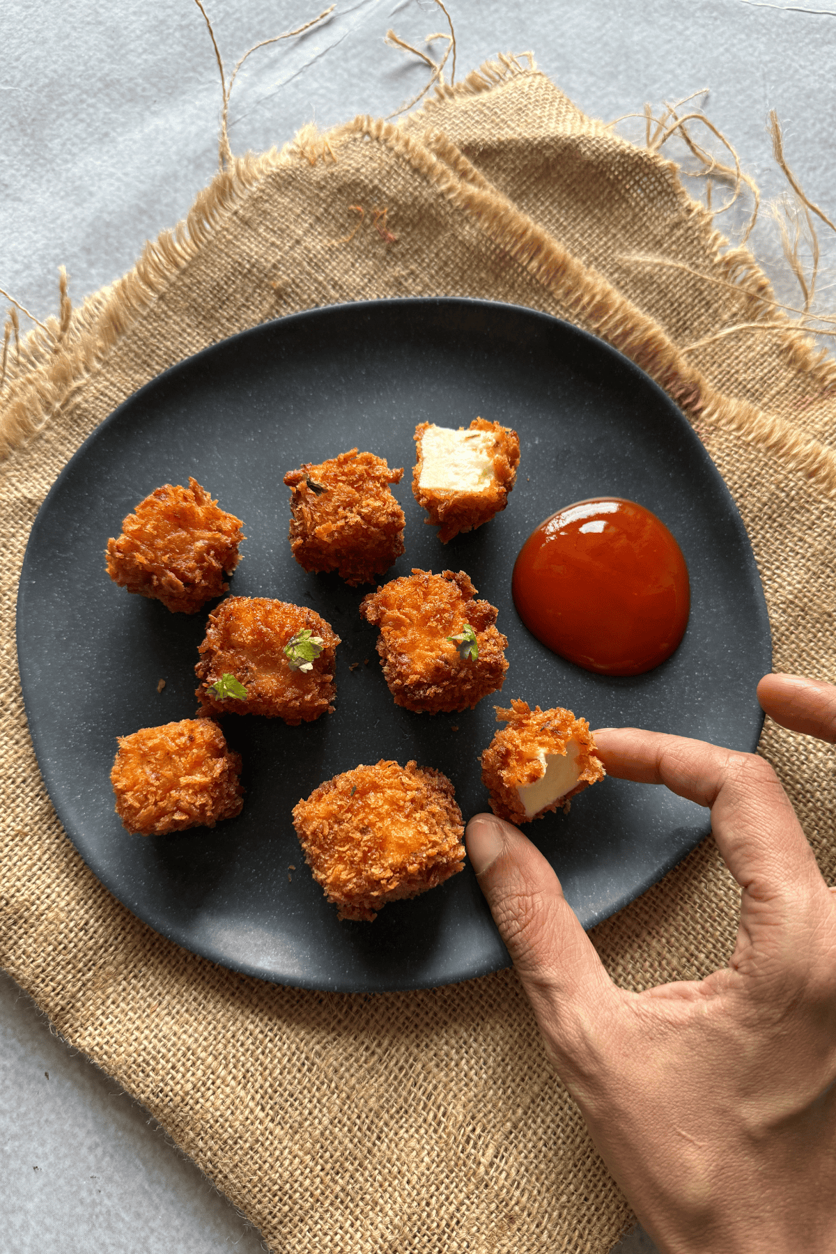 Crispy paneer popcorn pieces on a black plate with tomato ketchup, one piece held by hand showing the soft paneer inside, placed on a rustic burlap background.